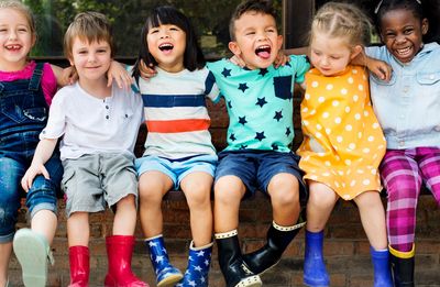 Six diverse young children sitting in a row laughing and smiling, wearing colorful clothing and rain boots